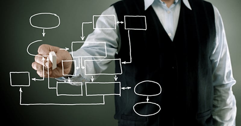 A man in a dark sweater vest draws a white flowchart of boxes, ovals and arrows against a dark background