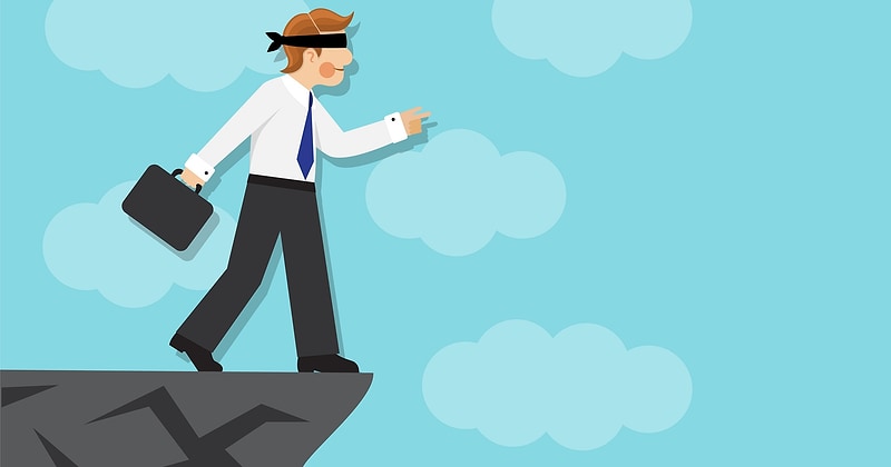 Flat illustration of a blindfolded businessman in a white shirt and blue tie, carrying a briefcase and reaching ahead with one hand as he steps to the very edge of a gray cliff above open sky